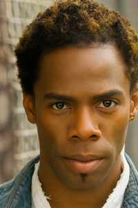 A head-and-shoulders portrait of a Black man with warm brown skin and short, tightly curled black hair featuring golden-blonde highlights. He has a small soul patch beneath his lower lip and wears a white shirt under a denim jacket. The man gazes directly at the camera with a serious expression. The background is softly blurred, showing a metal fence and neutral urban tones.